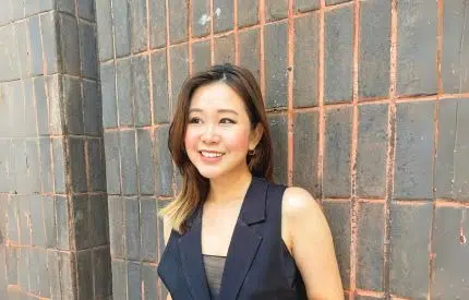 Melly Ling in a dark sleeveless blazer standing against a textured brick wall.