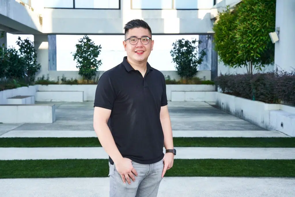 Melvin Chee in a black polo shirt standing outdoors in front of a modern building courtyard.