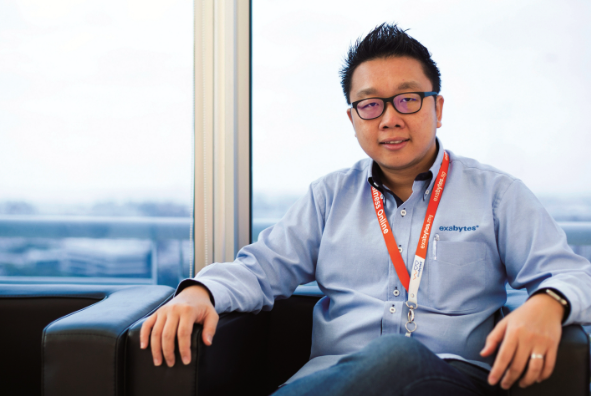 chan kee siak, CEO of Exabytes Network,  wearing glasses relaxes on a couch, positioned in front of a window that lets in bright sunlight.