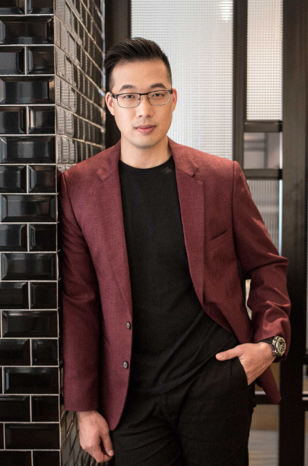 john son oei, founder and CEO of EPIC Collective, in a maroon blazer and black shirt leans casually against a textured brick wall.