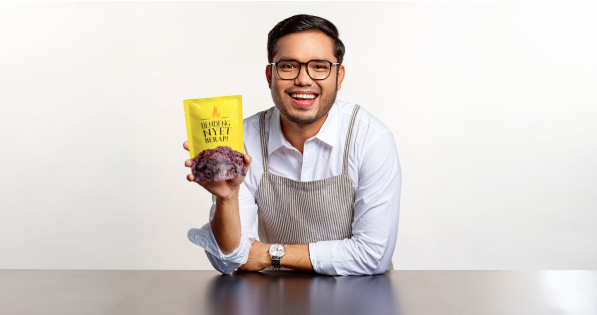 Khairul Aming wearing glasses holds a bag of food, smiling as he stands in a casual setting.